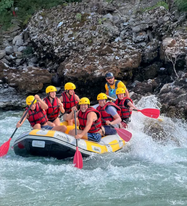 Seven rafters in red life jackets and yellow helmets paddling through white water below a rock wall on the Vjosa